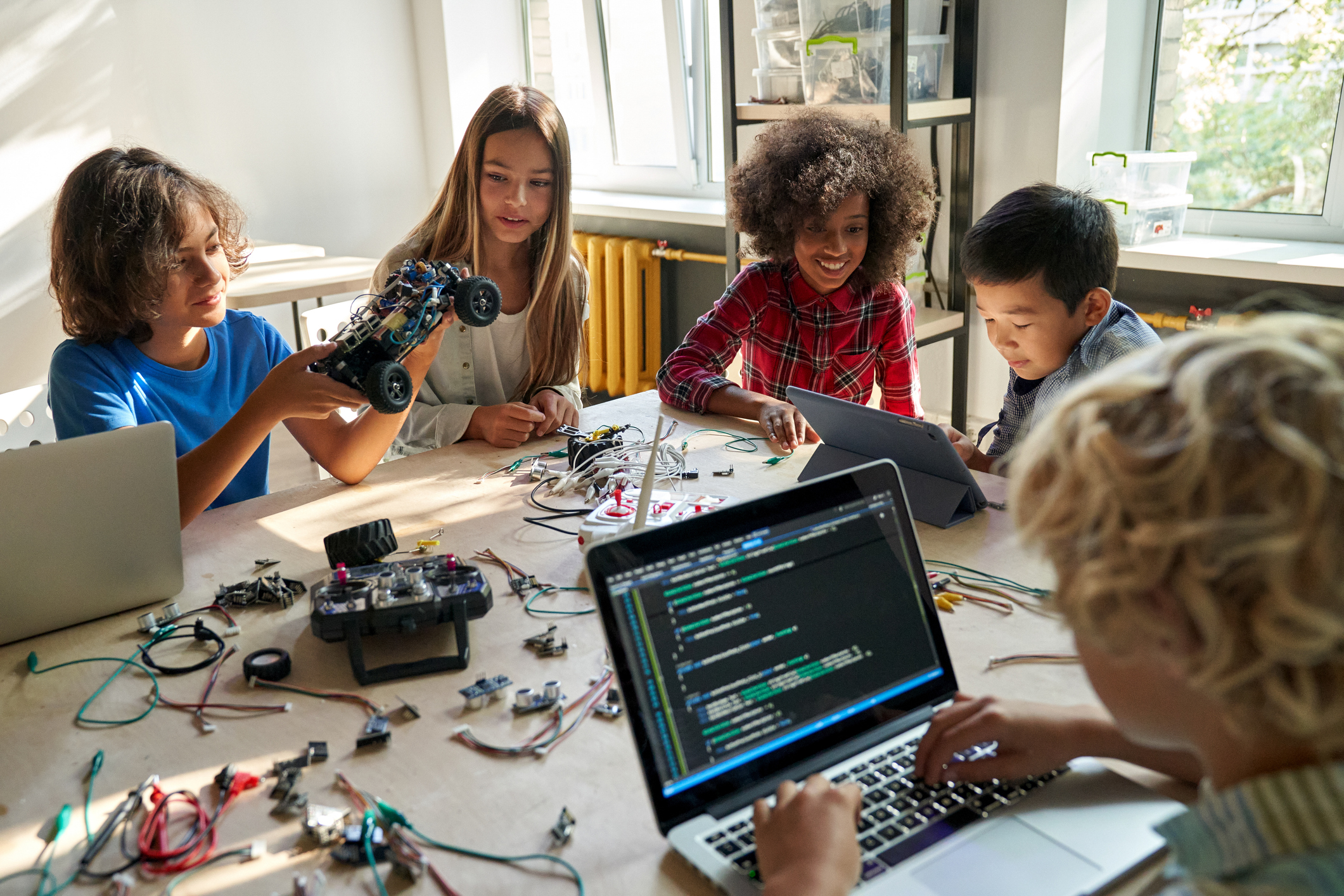 A group of teens building and testing an RC robot car with laptops and wiring spread across the table