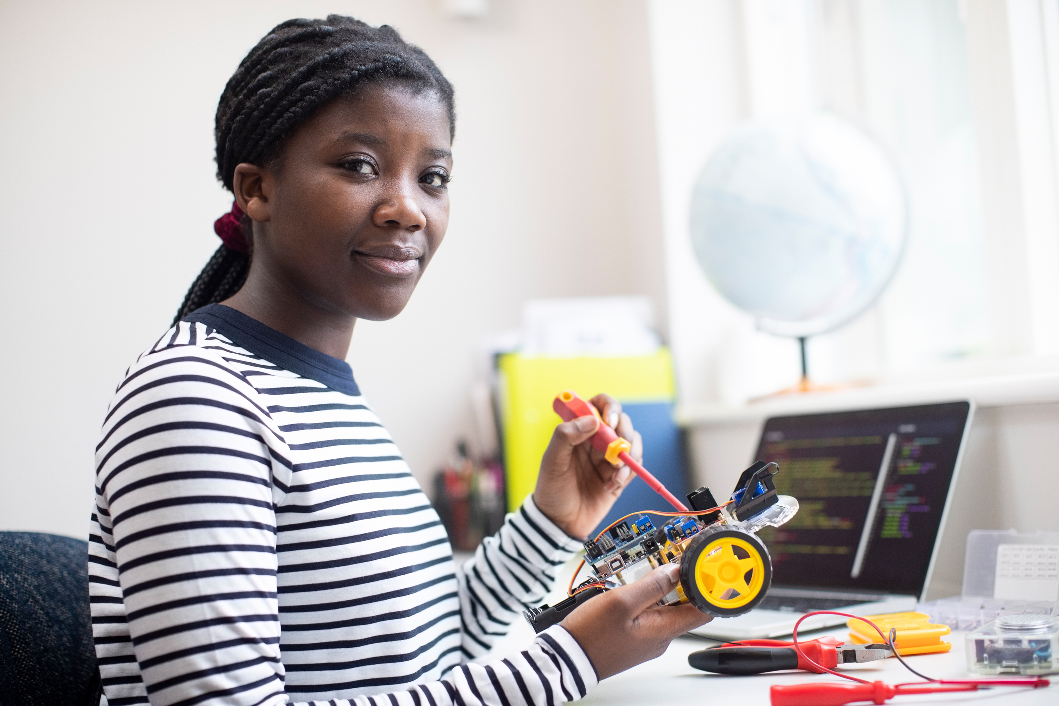 A smiling teen wiring a robot car with a screwdriver, laptop code visible on the screen behind her