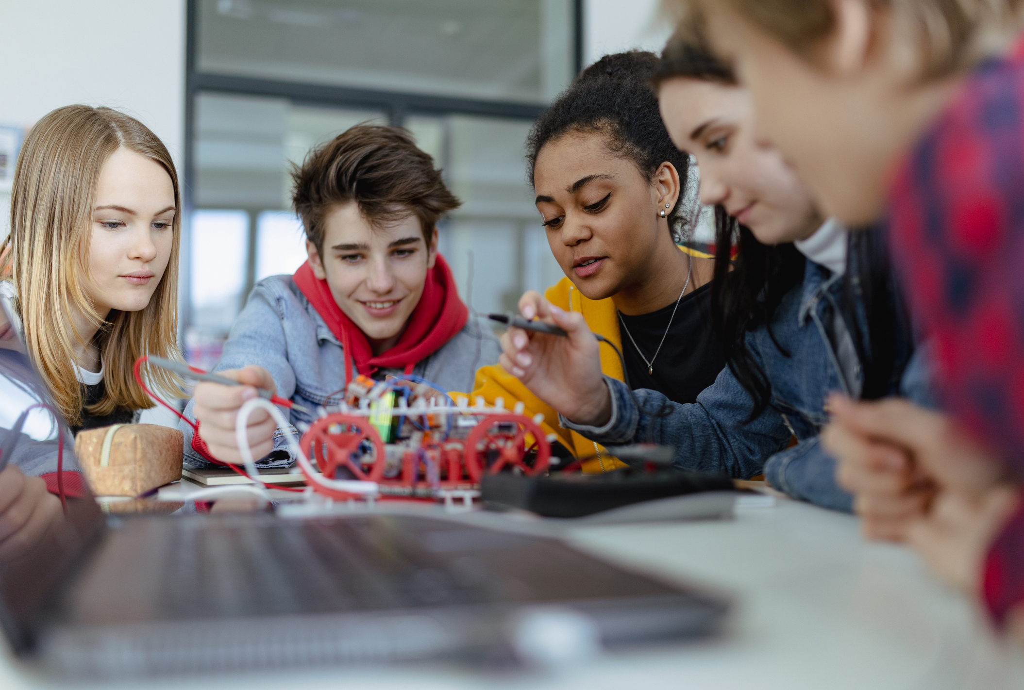 Five diverse teens leaning in together, wiring and assembling a robot car kit at a workbench