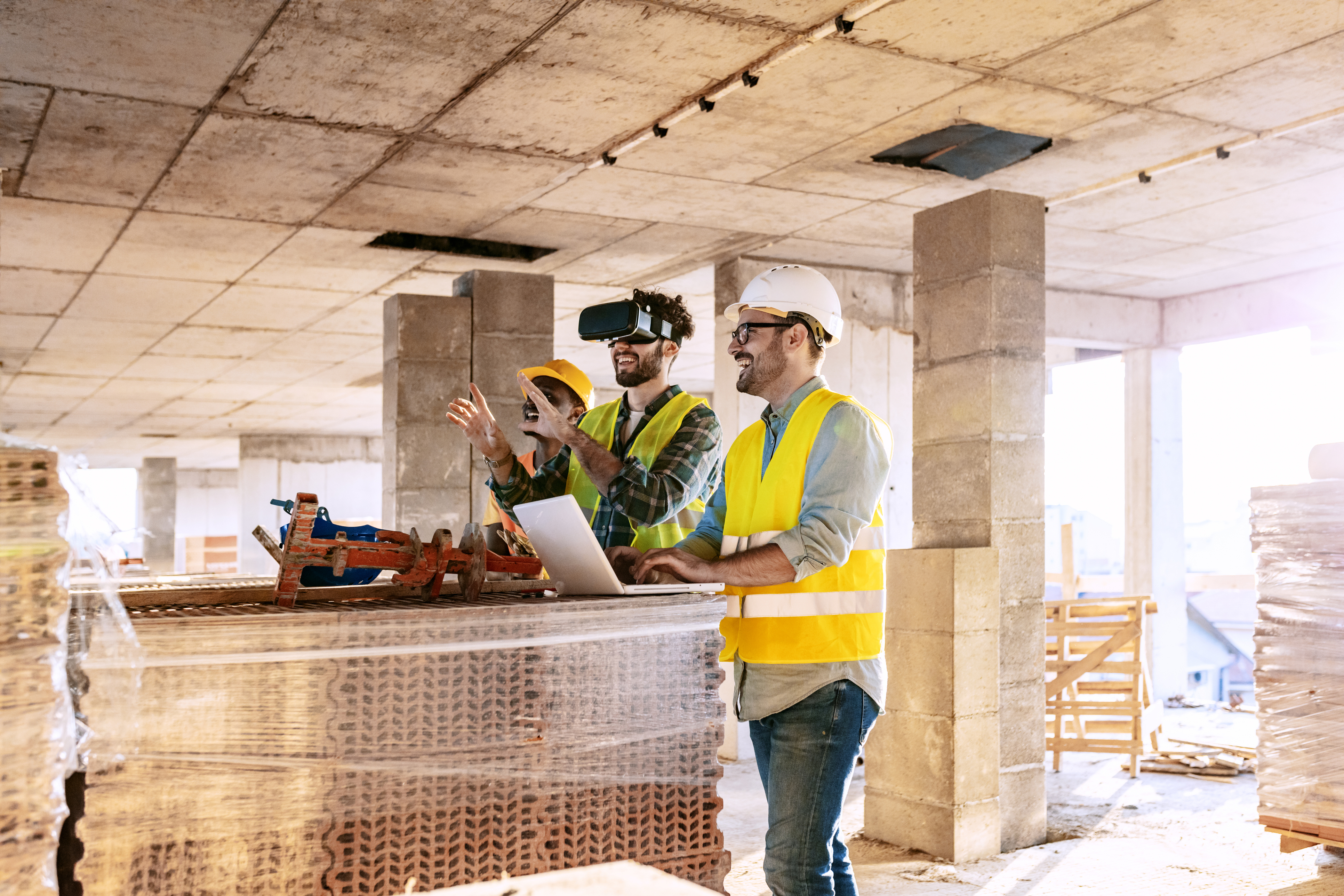 Construction workers in hard hats and hi-vis vests using a VR headset and laptop on an active building site